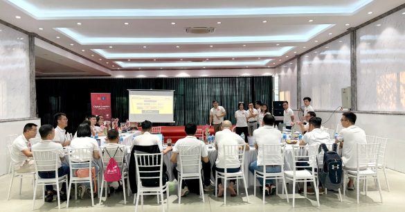 Lớp học viên Digital Leaders Coaching SS10 Hà Nội ngồi theo bàn tròn lắng nghe một nhóm thuyết trình bên màn chiếu và standee chương trình
