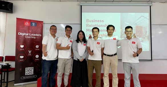 Nhóm sáu học viên Digital Leaders Coaching SS10 TP.HCM giơ ngón cái trước màn chiếu Business Relationship Management & Leadership, bên standee chương trình